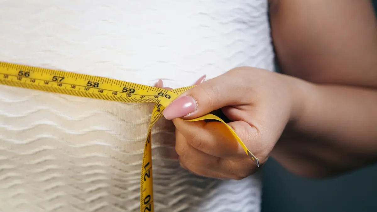 A person measuring their waist with a yellow tape measure against a textured white textile.