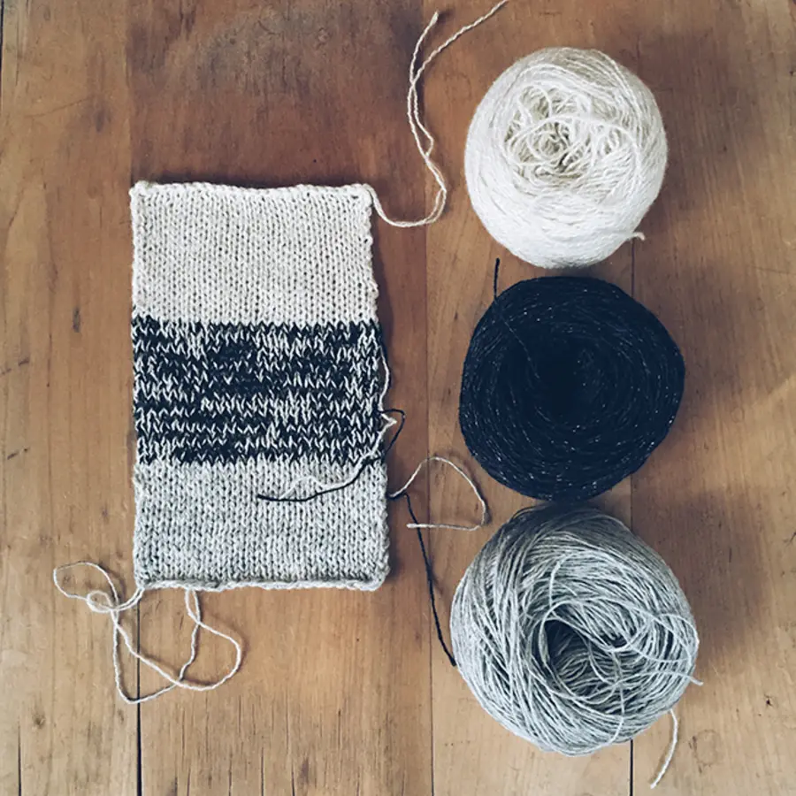 Knitted swatch with white, black, and gray sections beside three matching yarn balls on a wooden surface.