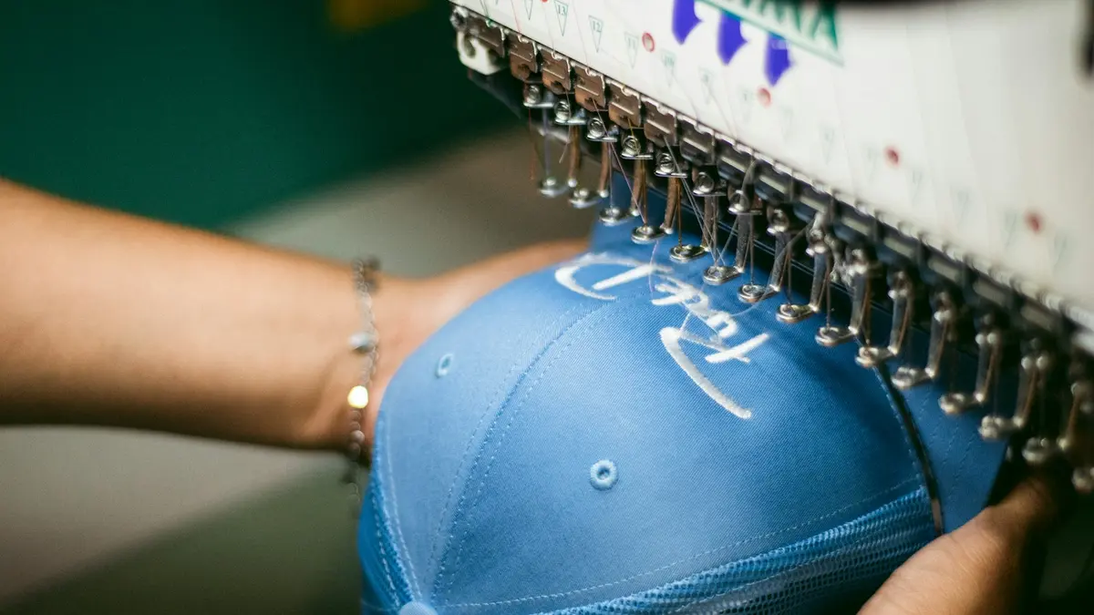 A blue cap is being embroidered with white thread by a machine.