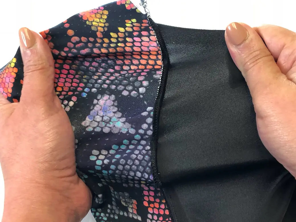 Hands stretch colorful patterned swimsuit fabric beside a smooth black panel, showing a low-bulk seam.