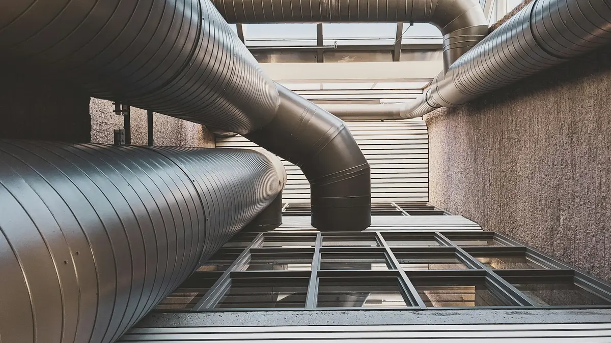 Metal pipes and ducts viewed from below, enclosed by textured walls and windows.