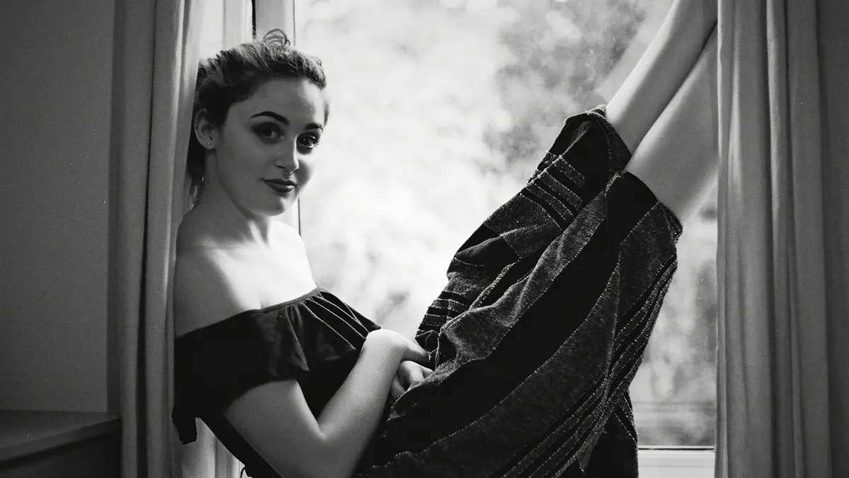 A woman in an off-shoulder top sits with her legs against a window, illuminated by diffused lighting.