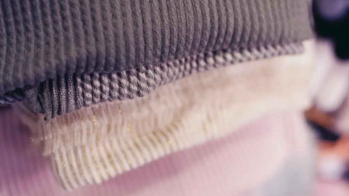 Stacked textiles in varying textures and colors, including grey, white, and pink.