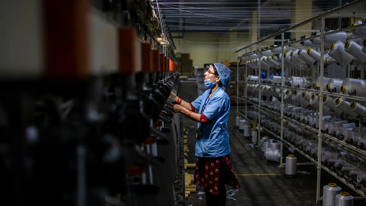 A woman operates a textile loom in a dimly lit factory, surrounded by spools of yarn.