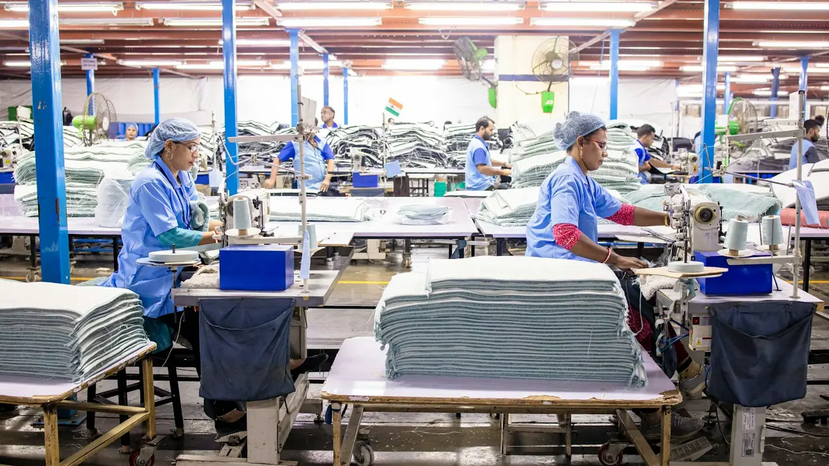 Workers operate sewing machines in a garment manufacturing facility with stacked fabric on tables.