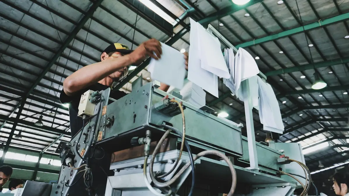 Worker operating a textile dyeing machine with sheets of paper in a factory setting.