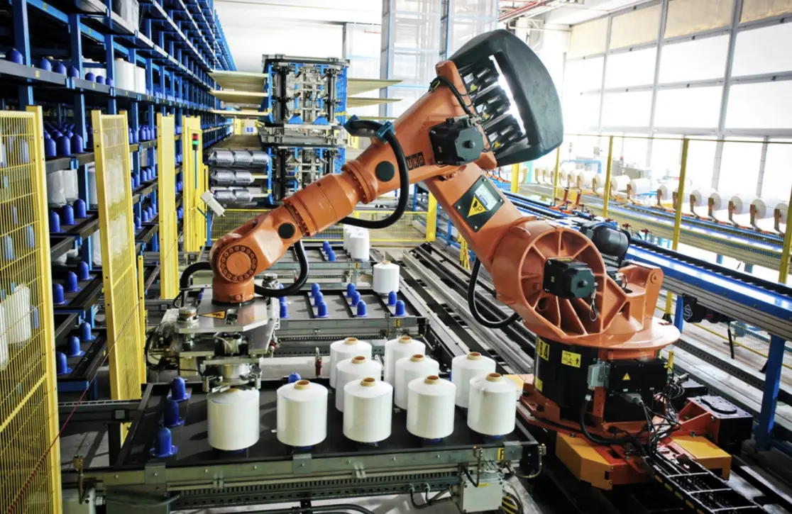 Industrial robotic arms handle white fabric rolls on a conveyor in a textile manufacturing facility.