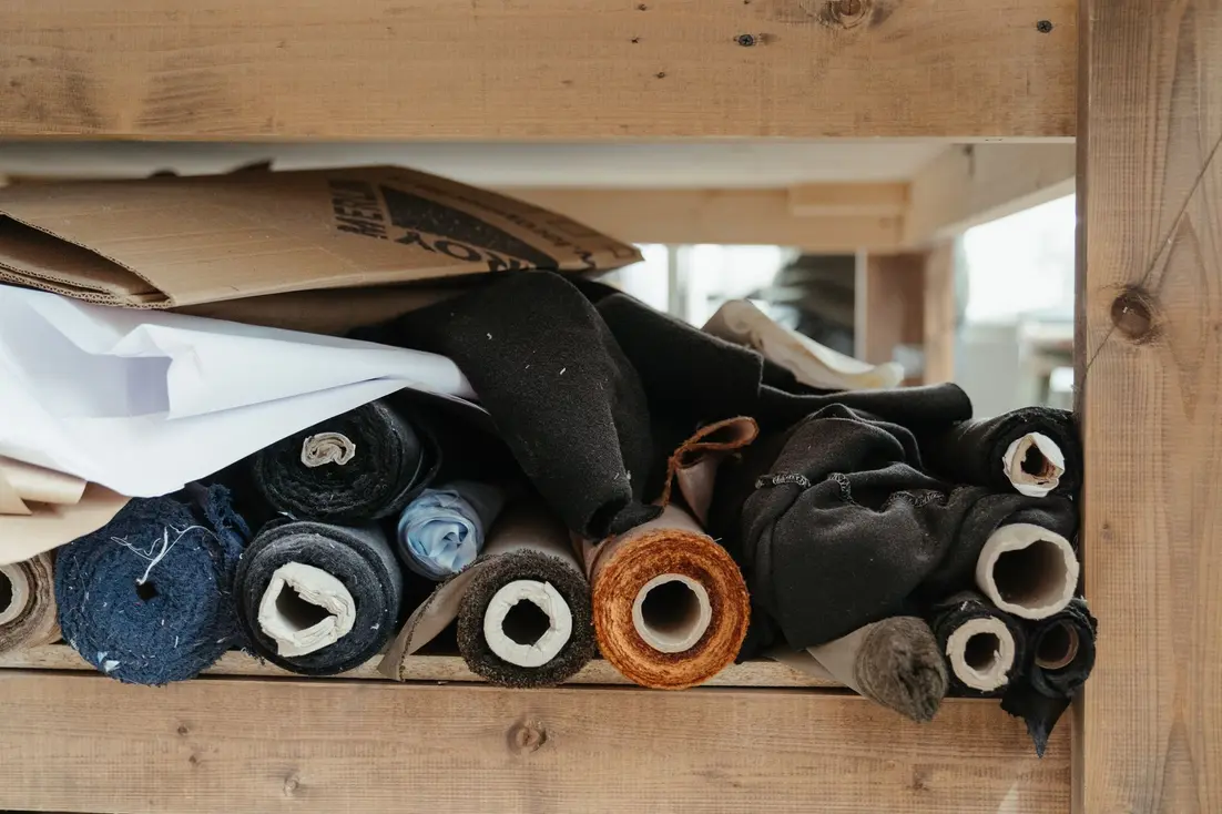 Rolls of various textiles neatly organized on a wooden shelf in a studio.